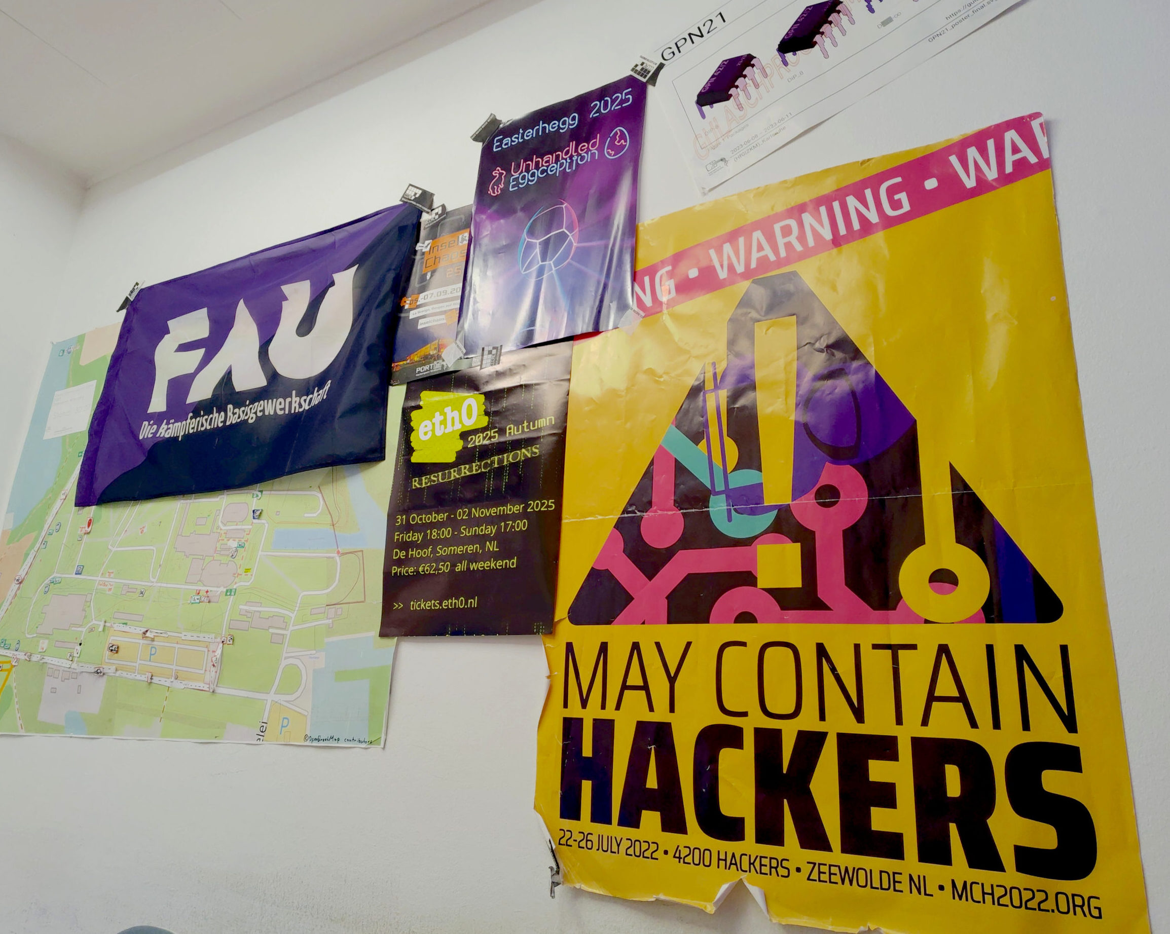 Mehrere Hackspace-bezogene Poster an einer weißen Wand. Eine FAU Fahne daneben.
